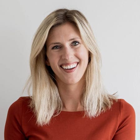 Vrouw met een vriendelijke glimlach en halflang, blond haar, gekleed in een oranje jurk. De afbeelding straalt een positieve en benaderbare uitstraling uit.