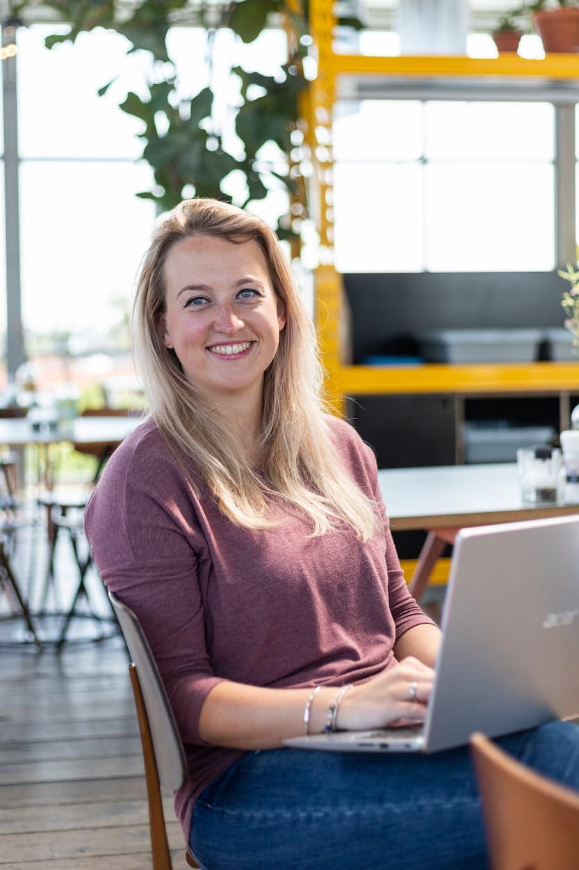 Vrouw met lang, blond haar zit in een moderne, lichte ruimte en werkt op een laptop. De achtergrond bevat groene planten en een stijlvol interieur, wat een inspirerende werkomgeving suggereert.