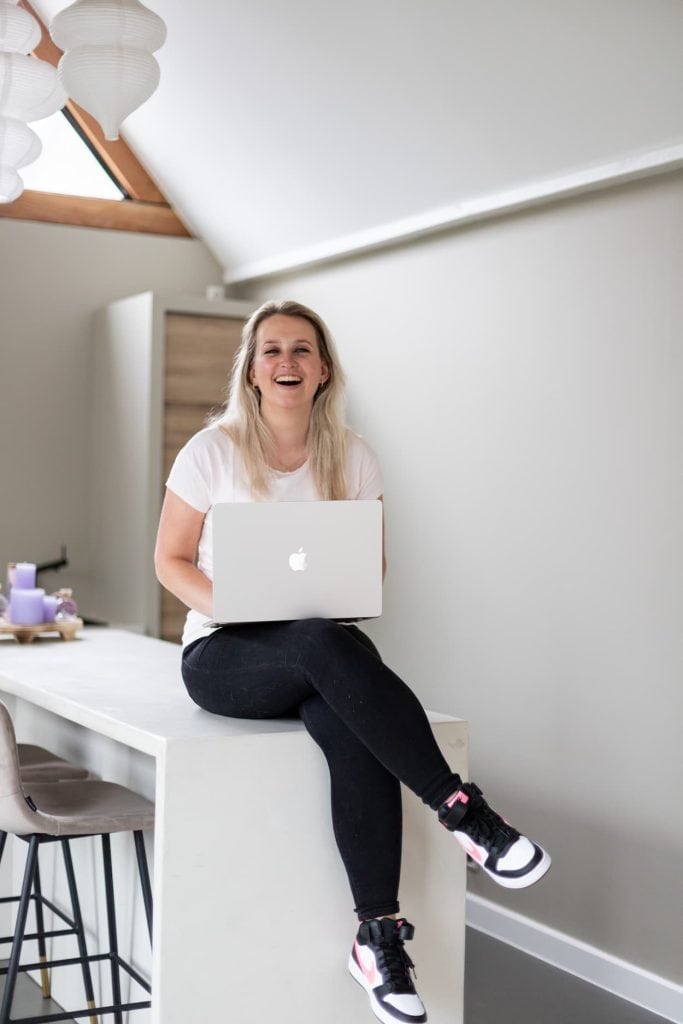 Een vrouw met lang, blond haar lacht terwijl ze op een moderne tafel zit en een laptop vasthoudt. De achtergrond heeft een minimalistisch ontwerp met een paar lichten en een neutrale kleurstelling.