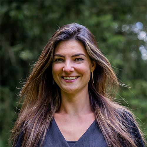 Professionele vrouw met lange haren en een vriendelijke glimlach, staand in een groene omgeving. De achtergrond bestaat uit bomen, wat een natuurlijke en ontspannen sfeer creëert.