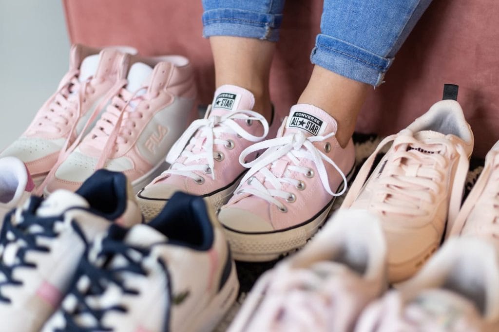 Roze sneakers van Converse All Star worden gedragen door een vrouw, omringd door verschillende andere sportschoenen. De focus ligt op de trendy en casual uitstraling van de schoenen.