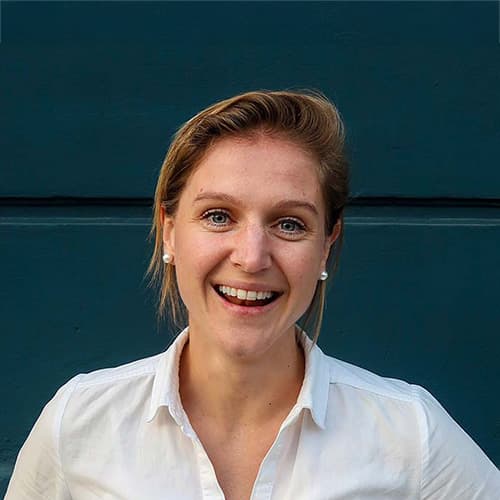 Vrouw met een glimlach, gekleed in een witte blouse, poseert voor een groene muur. Haar uitstraling straalt vriendelijkheid en enthousiasme uit.