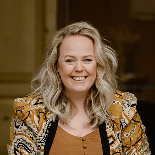 Vrouw met lang, golvend haar en een vrolijke uitstraling, gekleed in een kleurrijke, opvallende blazer. De achtergrond is neutraal en zorgt ervoor dat de focus op haar gezicht ligt.