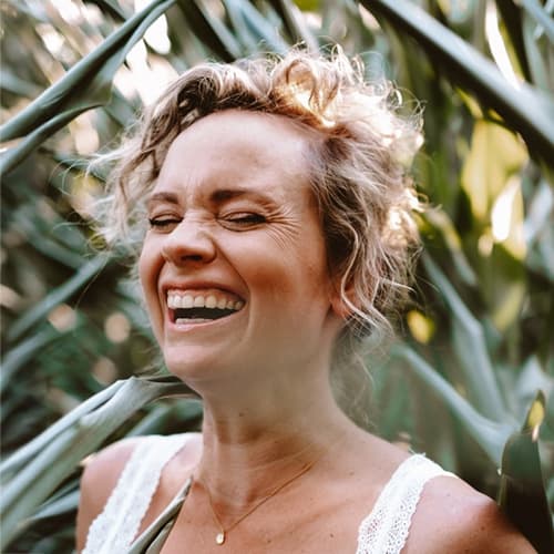 Een vrouw met een blije lach staat tussen groene bladeren, wat een gevoel van vreugde en natuur uitstraalt. Haar krullende haar en stralende gezicht benadrukken een moment van geluk en verbondenheid met de omgeving.