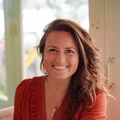 Een glimlachende vrouw met lang, golvend haar, gekleed in een oranje blouse met ruches, poseert vrolijk voor een lichte achtergrond. De afbeelding straalt warmte en vriendelijkheid uit.